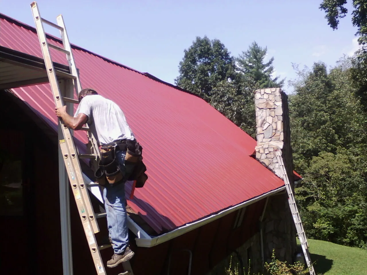 Roofer on a ladder beside a red metal roof and stone chimney — Soffit & Fascia Repair by Roofing Brothers in Barksdale